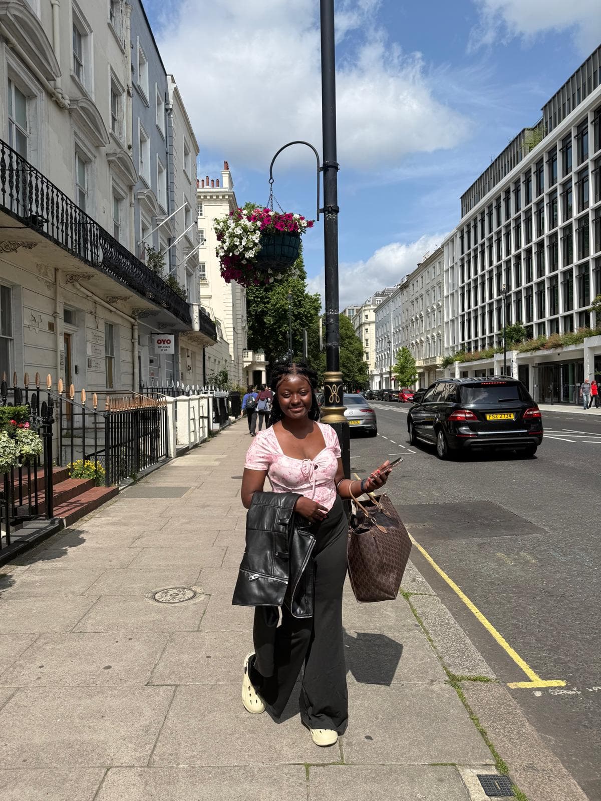 A young woman standing on a residential London street in summer, holding a phone and a bag, with white terraced houses behind her.