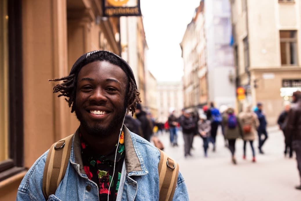 A smiling young man with a backpack standing on a busy European city street.