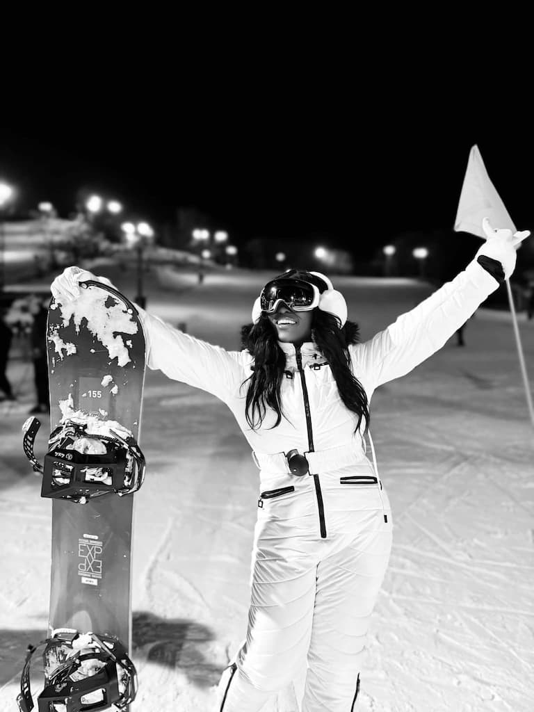 A black and white photograph of a woman in ski gear standing on a floodlit snow slope at night, holding a snowboard upright.