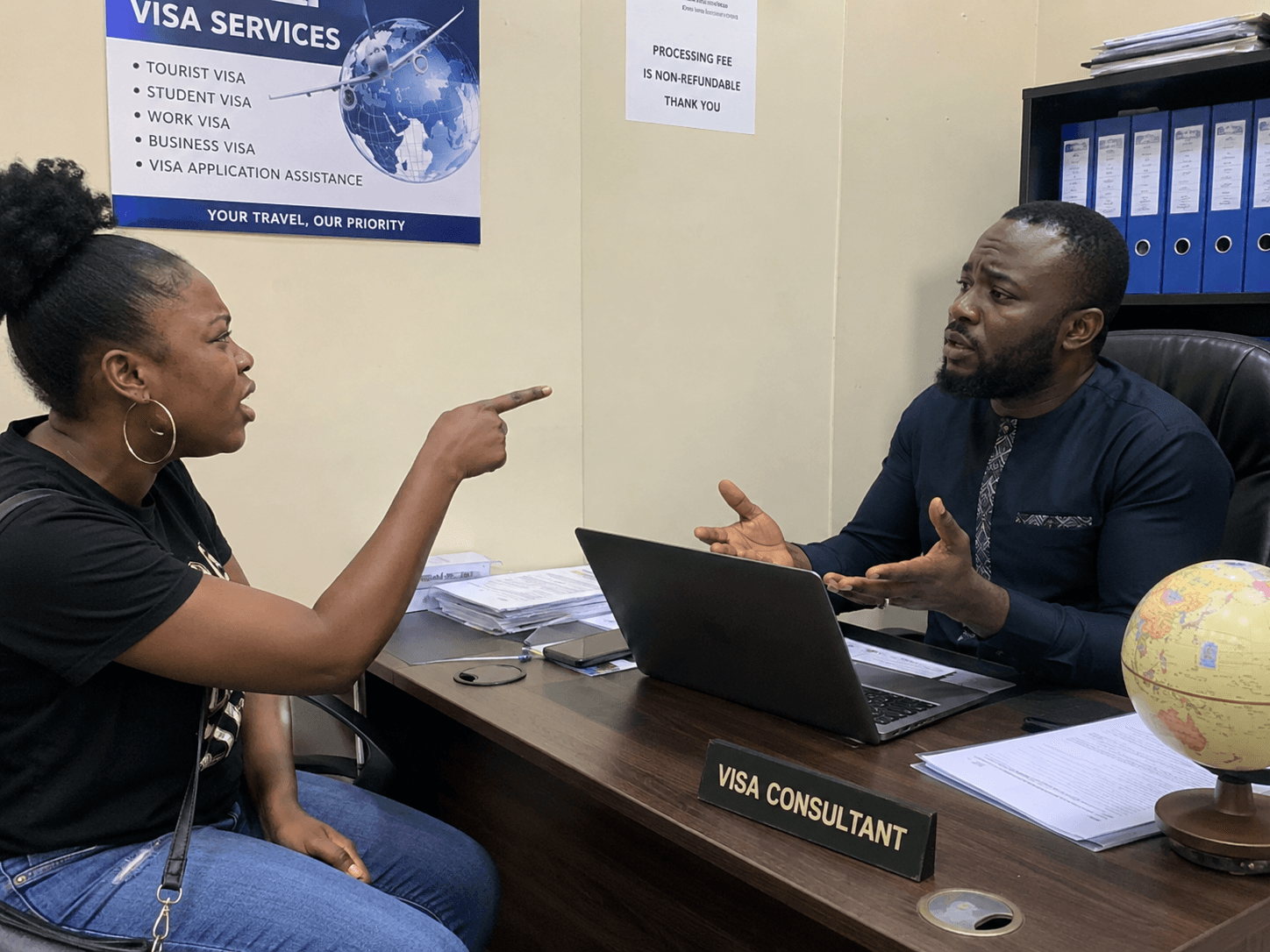 A woman gesturing in frustration across a desk towards a man seated behind a nameplate reading Visa Consultant, in an office with a visa services poster on the wall.